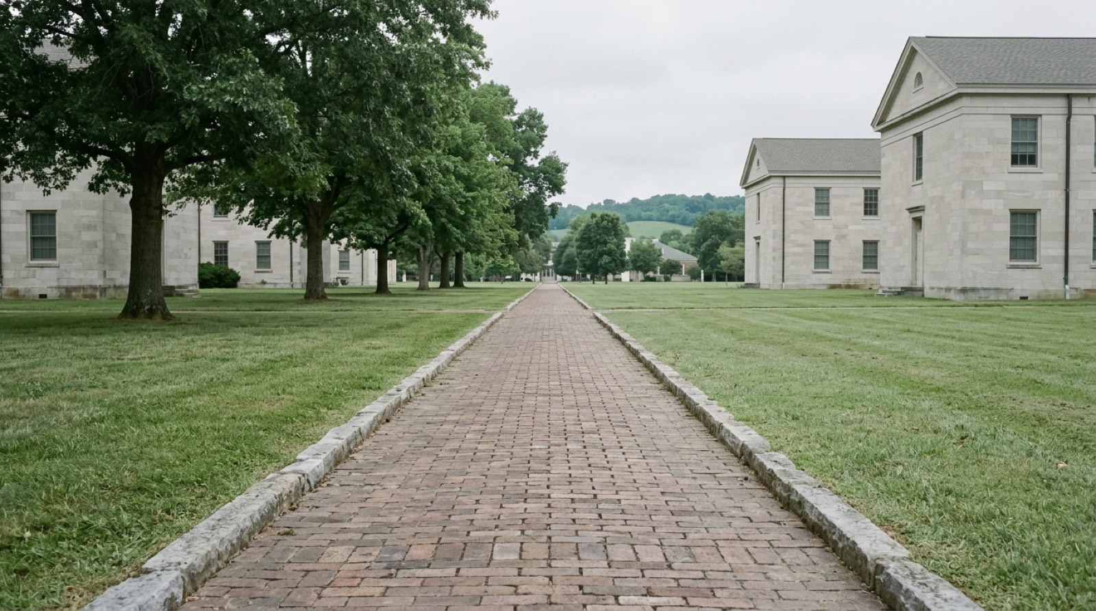 A brick footway running away between two plain limestone buildings across a mown quadrangle, with mature oaks along the left and low green hills behind, empty of people under a flat overcast sky